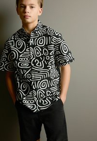 Kortärmad skjorta i svart med vita abstrakta mönster, knäppning framtill, kragdesign, i kombination med svarta shorts. Slät tygtextur.
