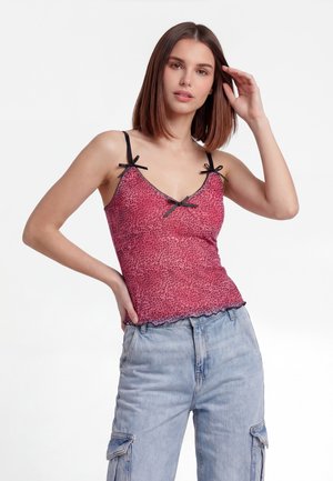 Junge Frau trägt ein rosa Leoparden-Top mit schwarzen Schleifen und hellblauen Cargohosen, posiert mit einer Hand auf der Hüfte und der anderen nahe am Kopf.