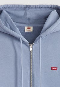 Felpa con zip azzurro chiaro realizzata in tessuto morbido. Presenta una zip frontale, cordini e un piccolo logo rosso di Levi's. Design a vestibilità standard.