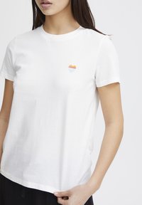 White cotton T-shirt featuring a small rainbow heart embroidered on the left chest, short sleeves, and a standard crew neck design.