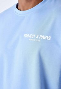 Lichtblauwe katoenen T-shirt met ronde hals en opvallende witte tekst: "PROJECT X PARIS MEMBERS' CLUB" gedrukt op de voorkant.