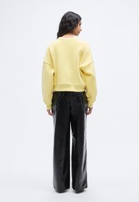 Κοντό κίτρινο φούτερ με χαμηλωμένους ώμους συνδυασμένο με μαύρο, ψηλόμεσο, γυαλιστερό παντελόνι με φαρδιά πόδια, από πίσω.