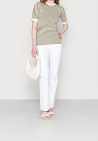 Mujer con una blusa de rayas en oliva y blanco de manga corta, pantalones blancos, sandalias trenzadas color crema, sosteniendo un pequeño bolso crema a su lado.