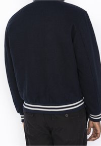 Giacca bomber blu navy con polsini e orlo a coste con strisce bianche e celesti. Il materiale sembra morbido e strutturato, senza loghi visibili.
