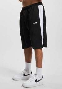 Shorts de sport noirs avec des bandes blanches sur les côtés, taille élastique et texte "DEF" brodé. Associés à des baskets blanches et des chaussettes.