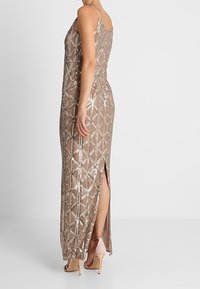 Abito senza maniche lungo fino al pavimento con motivo geometrico di paillettes, spacco laterale, indossato con sandali con tacco nudi, donna in piedi di lato.