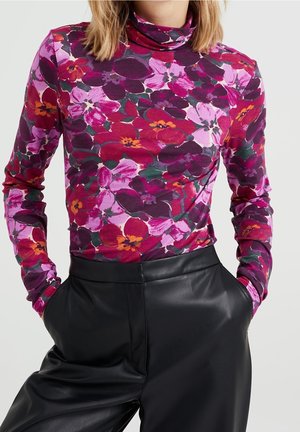 Kvinde iført en langærmet lilla, pink og rød blomster-rullekrave, der er stukket ned i højtaljede sorte læderbukser med hænderne i lommerne.