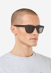 Ochelari de soare negri cu o finisare mată, ramă rectangulară și lentile întunecate, purtați de un model pe un fundal simplu.