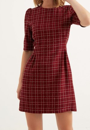 Femme portant une robe bordeaux courte, à manches courtes, avec un motif à carreaux blancs sur un fond uni.
