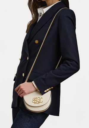 Sac crossbody en cuir crème avec ferrures dorées, présentant une forme incurvée et des initiales moulées "RBL", porté avec un blazer bleu marine à double boutonnage.