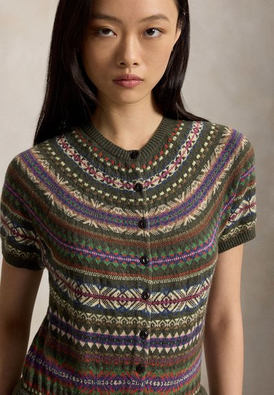 Jeune femme aux longs cheveux foncés portant un cardigan à manches courtes boutonné avec des motifs géométriques multicolores et fair isle.