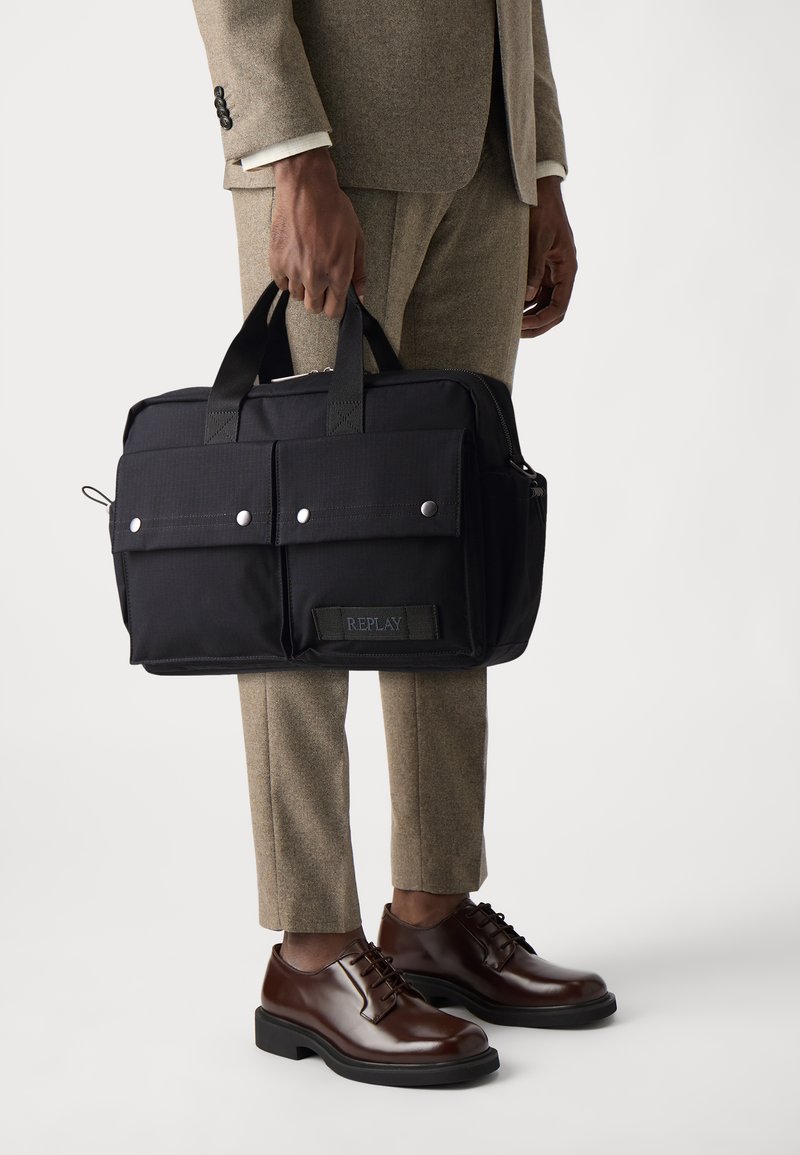 Sort nylon messenger bag med to frontlommer, sølv snaplukninger og REPLAY-branding. Holdt i en hånd, parret med brune sko.