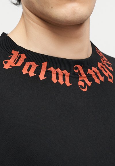 Κοντινό πλάνο ενός ατόμου που φοράει μαύρο μπλουζάκι με στρογγυλή λαιμόκοψη, με την επιγραφή "Palm Angels" σε κόκκινη γοτθική γραμματοσειρά γύρω από το γιακά.