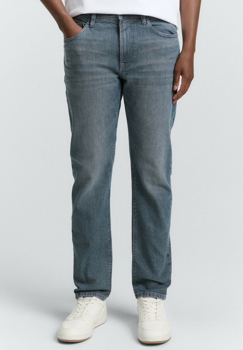 Person in blauen Jeans und weißen Sneakern, steht mit einer Hand in der Tasche vor einem schlichten Hintergrund.