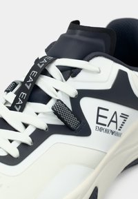 Fehér és tengerészkék sportcipő texturált szintetikus felsőrésszel, kerek fűzőkkel és "EA7" valamint "Emporio Armani" márkajelzésű címkével.