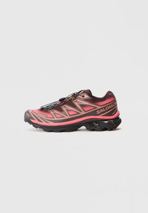 Zapatilla de trail running con un diseño degradado en marrón y rosa, que presenta una parte superior texturizada, una suela negra y un sistema de cordones único.