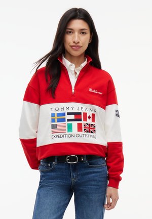Frau trägt ein rot-weißes Sweatshirt mit Viertel-Reißverschluss und mehreren Länderflaggen, "Tommy Jeans Expedition Outfitters" Text und blaue Jeans.