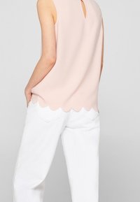 Kvinna som bär en ärmlös ljusrosa topp med vågig fåll och vita byxor, sedd bakifrån, med händerna i fickorna.