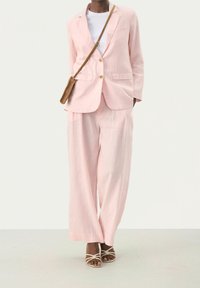 Blazer en lin rose clair à deux boutons, assorti d'un pantalon large de la même couleur. Porté avec un haut blanc et des sandales à brides beiges.