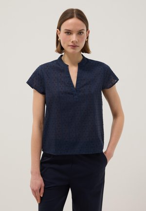 Femme aux cheveux bruns et raides, portant un chemisier bleu marine à manches courtes avec une texture subtilement à pois et un pantalon bleu marine assorti.