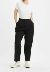 Pantaloni neri a vita alta con pieghe, realizzati in tessuto solido. Abbinati a una maglietta attillata bianca a collo rotondo e sneakers di colore chiaro.