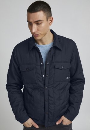 Marineblau gesteppte Jacke mit zwei Brusttaschen, Druckknöpfen und einem Kragen, über einem hellblauen Hemd geschichtet. Weiche Textur.