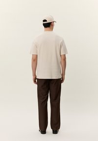 Vīrietis, valkājot bēša krāsas cepuri un T-kreklu ar brūniem biksēm, stāv ar muguru pret vienveidīgu gaišu fonu.