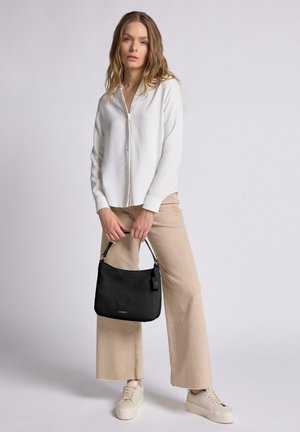Black leather handbag with textured surface, gold hardware, and a detachable tag, paired with a white blouse and beige wide-leg pants.