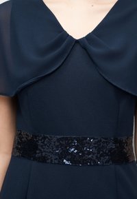 Ναυτικό μπλε φόρεμα με στρωματοποιημένη λαιμόκοψη V και φαρδιά μαύρη ζώνη με παγιέτες γύρω από τη μέση.