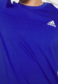 Sininen adidas t-paita, joka on valmistettu pehmeästä kankaasta. Paita sisältää pyöreän kaula-aukon, lyhyet hihat ja valkoisen logon vasemmassa ylärinnassa.