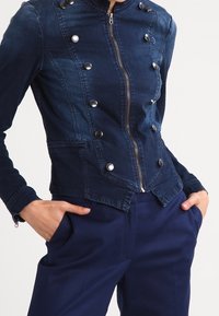 Chaqueta de denim en azul oscuro con un diseño ajustado, cuello alto, botones de presión plateados y cierre con cremallera. Presenta bolsillos laterales y costuras ajustadas.