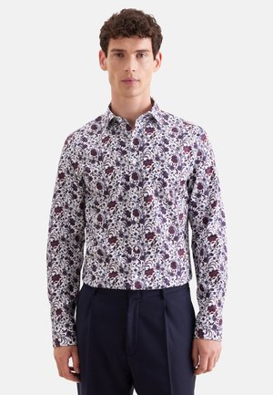 Mann trägt ein langärmliges Hemd mit weißem, lila und rotem Blumenmuster und dunkelblauen Hose.