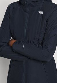 Granatowa wodoodporna kurtka z zamkiem błyskawicznym, kapturem i elastycznymi mankietami. Posiada logo na klatce piersiowej i rękawie. Gładka tekstura.