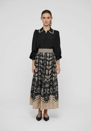 Kvinde stående iført en sort bluse med lange ærmer med mønstret krave og sort og beige blomster midi nederdel med sorte spidse sko.