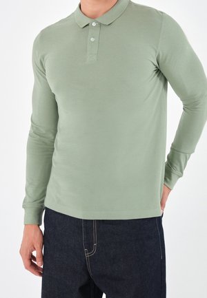 Langärmliges Polo-Shirt aus leichtem grünem Baumwollstoff, mit Kragen, drei Knöpfen und einer taillierten Passform, kombiniert mit dunklen Jeans.