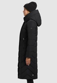 Vrouw in een lange zwarte gewatteerde jas met capuchon en zwarte gebreide beanie, staand in profiel tegen een effen lichte achtergrond.