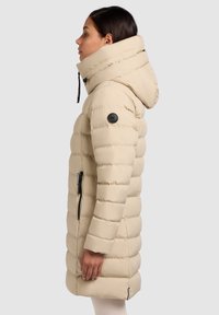 Beežikas pufferjope vertikaalsete segmentide, kõrge krae ja reguleeritava kapuutsiga. Varustatud lukuga taskute ja vasakul varrukal musta logomärgiga.