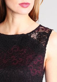 Svart och bourgogne spetsöverdel med ett blommönster, med rundad halsringning och texturerade detaljer på tyget.