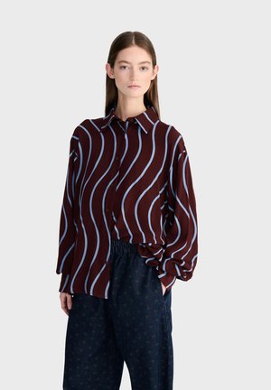 Jeune femme debout les mains dans les poches, portant un chemisier bordeaux à rayures ondulées bleu clair et un pantalon ample foncé à motifs.