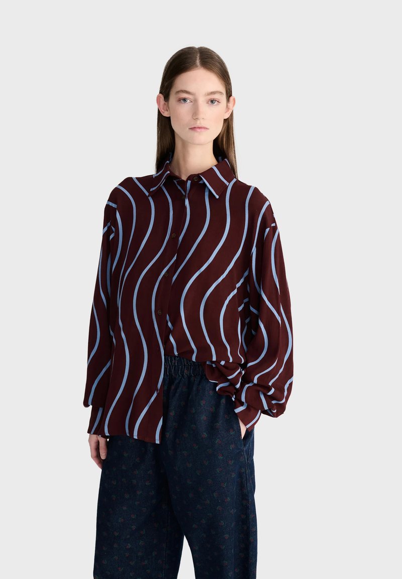 Jeune femme debout les mains dans les poches, portant un chemisier bordeaux à rayures ondulées bleu clair et un pantalon ample foncé à motifs.