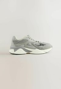Chaussure de sport grise présentant un mélange de daim texturé et de mesh, avec une semelle contrastante blanche et gris clair. Conçue avec des lignes géométriques bien marquées.