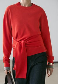 Röd tröja med knytbar midja, ribbad halsringning och muddar, kombinerad med mörka jeansbyxor. Mjuk, len tygstruktur.