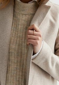 Beige villakangas takki vaalean kermaisen ryppyisen pooloneuleen päällä, käsi pitämässä takin kaulusta. Tekstuurit eroavat hienovaraisesti.