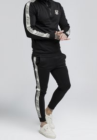 Black athletic tracksuit set with quarter-zip top, featuring white patterned stripes on sleeves and pants, paired with white sneakers.