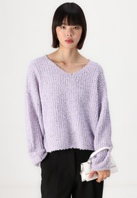Suéter de malha lavanda com decote em V e corte solto. Tecido texturizado e mangas longas, combinado com uma pequena bolsa branca.