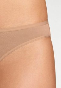 Perizoma beige senza cuciture con una texture liscia, cintura elastica e copertura minima, progettato per comfort e discrezione sotto i vestiti.