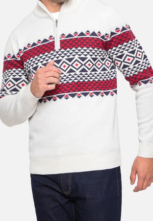Homme portant un pull en maille blanche avec un motif géométrique rouge, noir et blanc sur la poitrine et le haut des bras, associé à un jean bleu foncé.