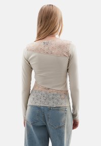 Langarmshirt in Beige mit Spitze an Schultern und Rücken, mit taillierter Passform und weichem Material, kombiniert mit hochgeschnittenen Jeans.