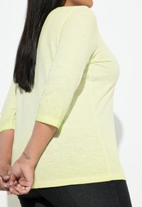 Jasnożółta bluzka z długim rękawem, okrągłym dekoltem i luźnym krojem, wykonana z lekko teksturowanego materiału, z kontrastowym obszyciem na rękawach.