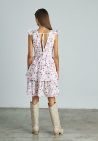Weißes Blumenkleid mit Rüschärmeln und einem gestuften Rock. Verfügt über einen tiefen Ausschnitt im Rücken mit einem Band zum Binden. Kombiniert mit beigen knielangen Stiefeln.